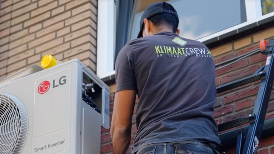 Monteur van KlimaatCREW op een ladder bij een buitenunit van een airco tegen een gevel